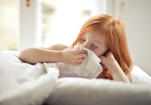 Redheaded child resting in bed, wiping nose with tissue, unwell with flu-like symptoms.